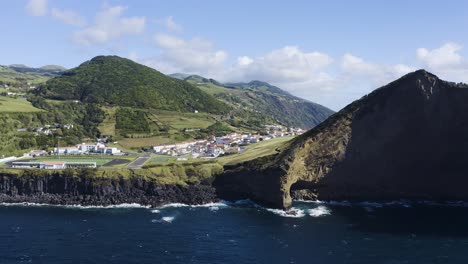 eingestürzte vulkan-caldera, die ein dorf, einen skatepark, einen sportplatz mit bergen, vegetation und lavaformationen neben dem meer freigibt. drohnenaufnahmen, velas, insel sao jorge, azoren, portugal