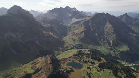 A-stunning-aerial-view-of-Warth,-Vorarlberg,-Austria,-with-the-majestic-Alps-as-a-backdrop,-sunlit-hills,-and-a-hazy-sky-dotted-with-light-clouds-on-the-horizon