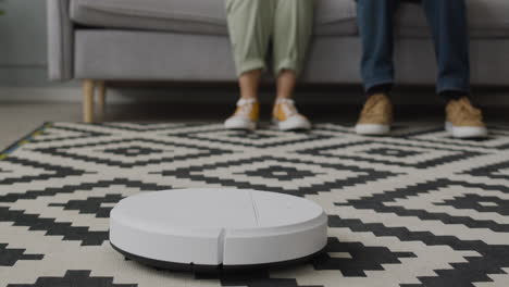 close up of a robot vacuum cleaner tidying the room while two unrecognizable people sitting on sofa at home