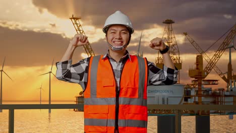 ingeniero asiático con casco de seguridad flexionando su bíceps y sonriendo a la cámara mientras está de pie con la producción de hidrógeno en alta mar