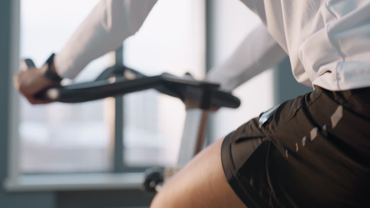 close up of man gripping handlebars of stationary bike in bright gym light filtering through window as he glances away, suggesting momentary distraction during indoor cycling workout session