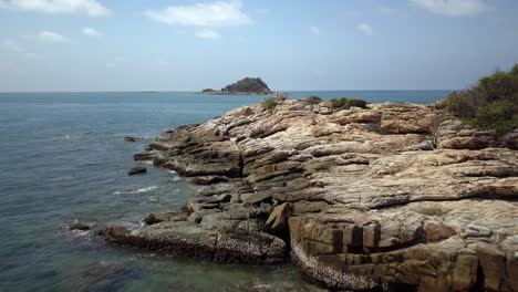 aerial: a low altitude , close to the water forward shot of the sharp stones and a rocky shore, the drone rises up over it and reveals a small island in the distance