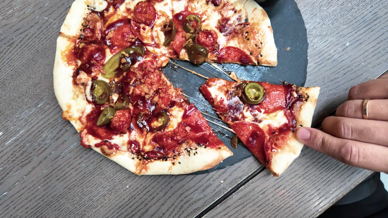 Un primo piano mostra una mano che solleva una fetta di pizza al formaggio, pepperoni e jalapeño da una pizza rotonda rustica su un tavolo di legno, con formaggio fuso e condimenti vivaci alla luce naturale del giorno