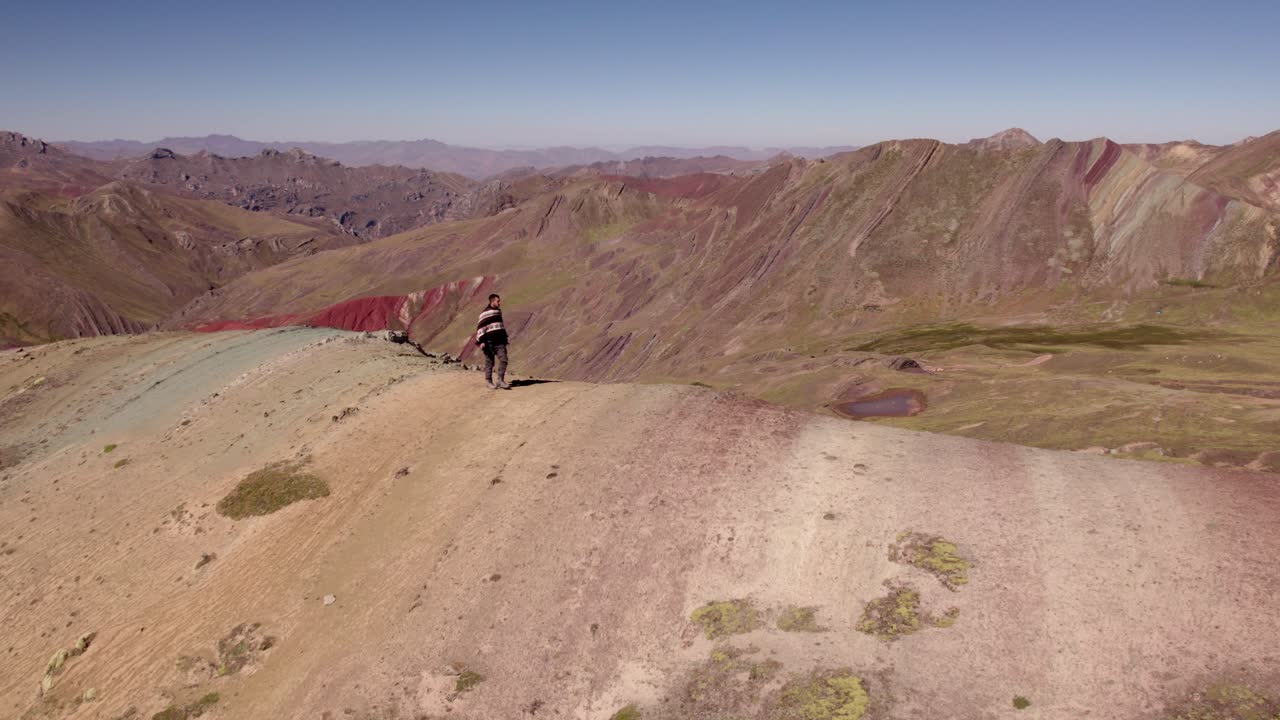 A hiker traverses a colorful ridge in the Peruvian Andes, showcasing stunning mineral hues. The expansive landscape features rolling hills and endless mountain ranges under clear skies.