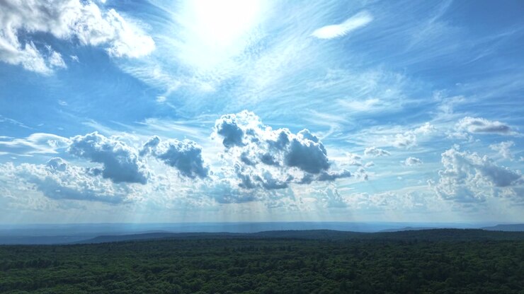 Las imágenes aéreas capturan rayos de sol que se filtran a través de nubes dramáticas sobre un vasto bosque verde. La toma de gran angular resalta el cielo azul vibrante, las nubes texturizadas y el sereno paisaje natural debajo.