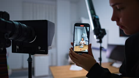 female hands hold the smartphone taking video of a lady sitting at desk. girls records a speech of blogger in studio. blurred backdrop.