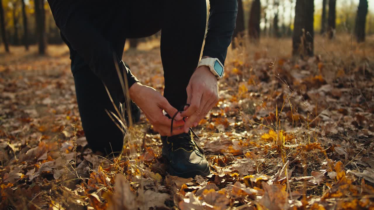 cerrar un hombre en un uniforme deportivo negro ata los cordones de sus zapatillas antes de comenzar su carrera a través del bosque soleado de otoño por la mañana. hombre confiado con el cabello rizado comienza su carrera por la mañana en el bosque