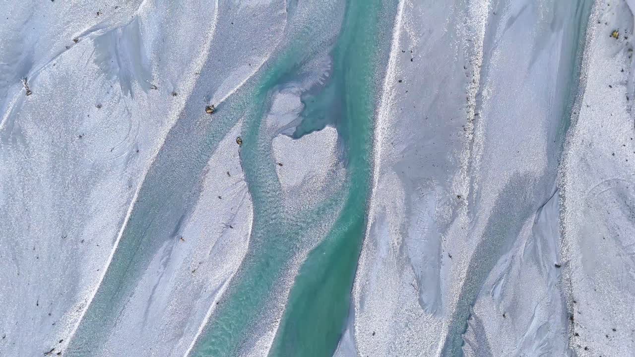 Aerial footage captures the intricate patterns of a turquoise braided river flowing through a rocky landscape in Glenorchy, New Zealand