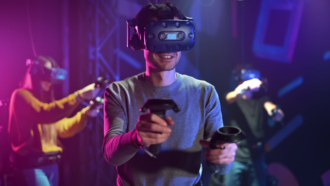 A joyful, interested man is playing on VR treadmill with helmet and controllers. Other people playing nearby