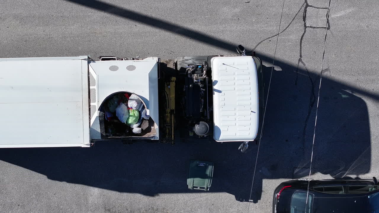 Static top down aerial drone footage where a dump truck drives into view and picks up a trash can to dump.