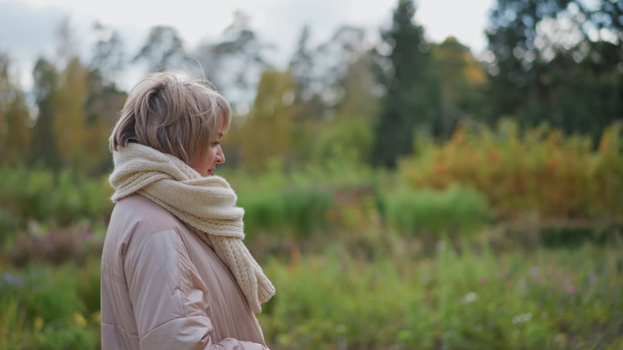 close up side view of woman in business coat and chunky neck muffler strolling reflectively through lush green garden path lined with tall trees and blurred foliage
