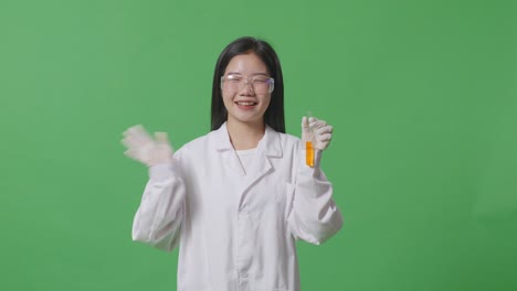 asian woman scientist with orange liquid in the test tube smiling and waving her hand while standing on the green screen background in the laboratory