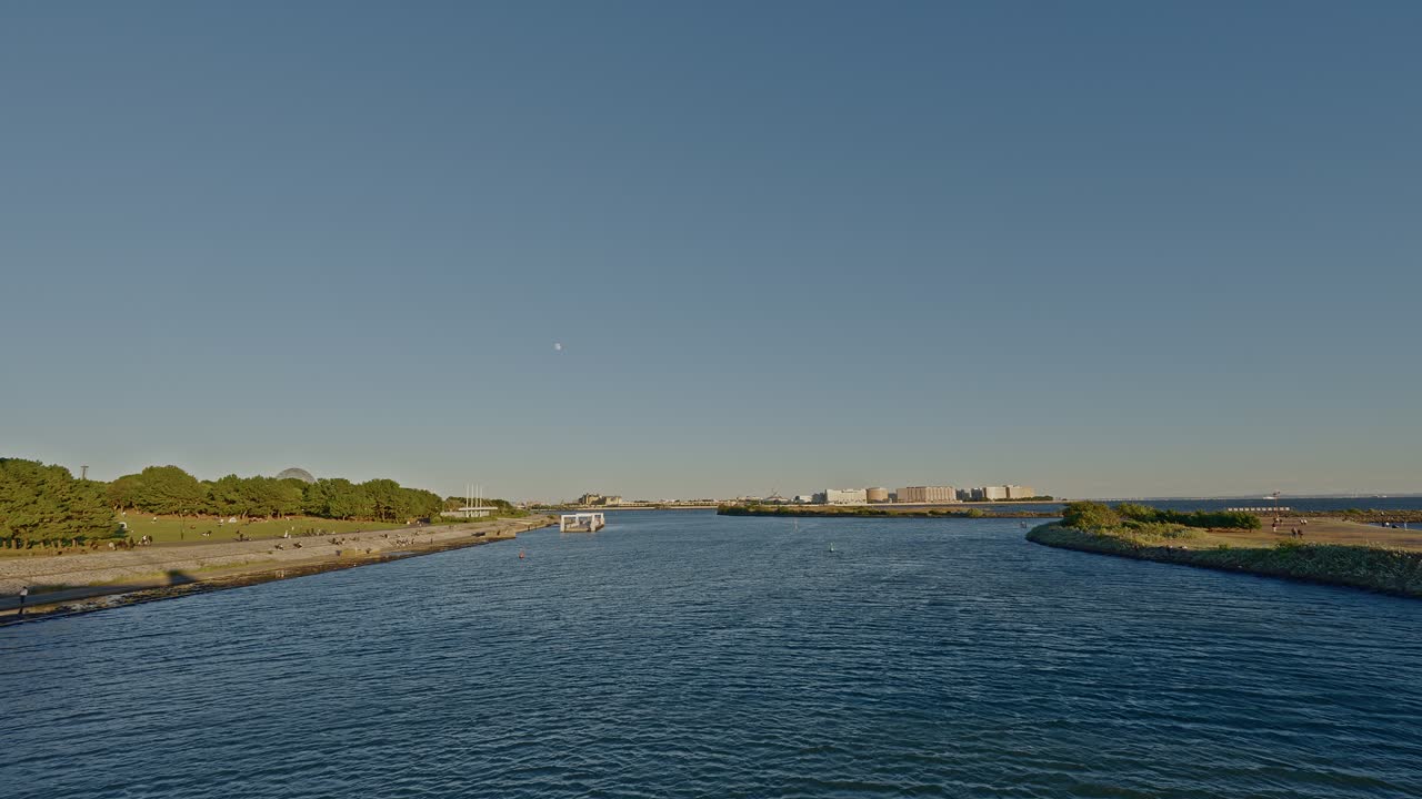 Wide shot of a peaceful, sunlit river or harbor channel with a small boat traveling toward a distant cluster of large buildings