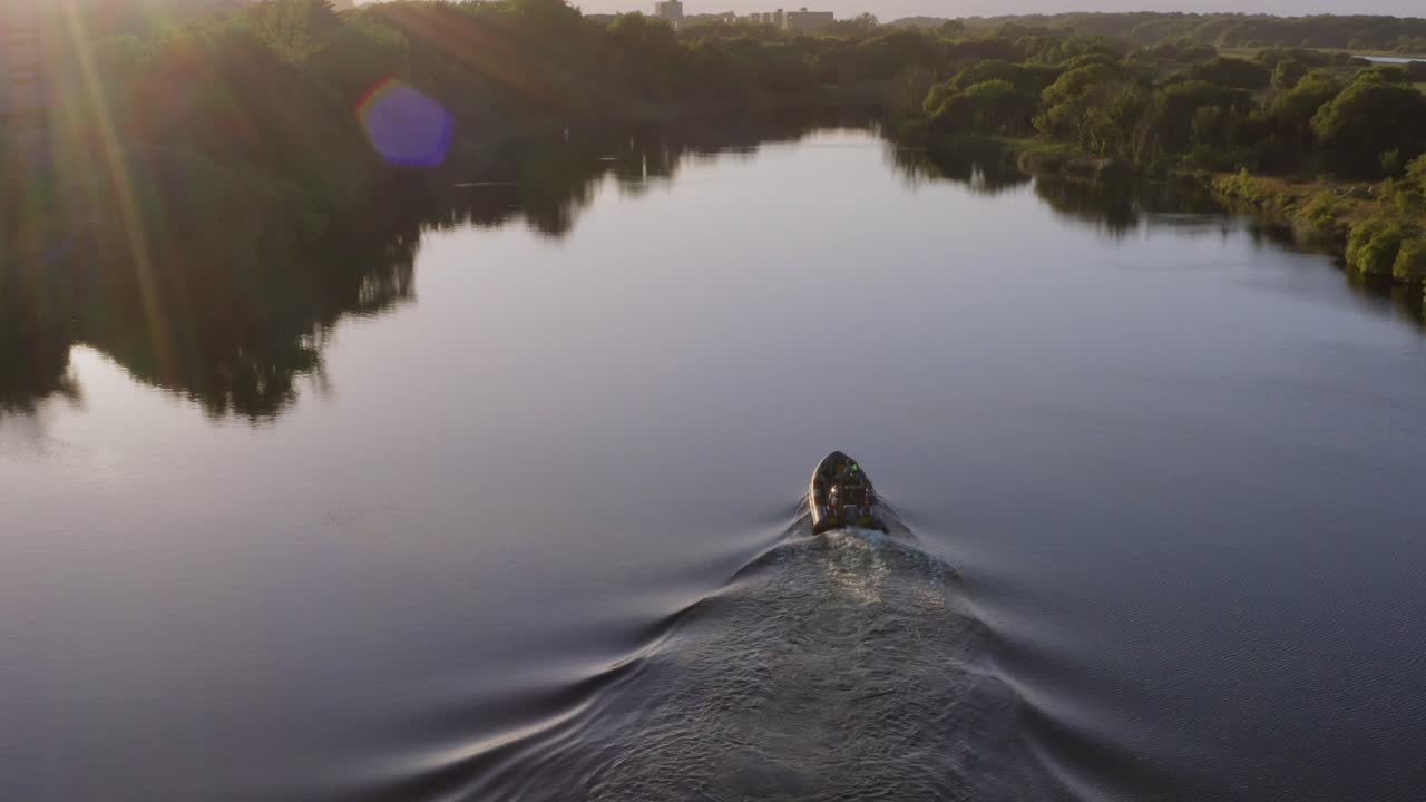 Inflatable pontoon boat drives up river as sun flare shines down and wake breaks calm water surface