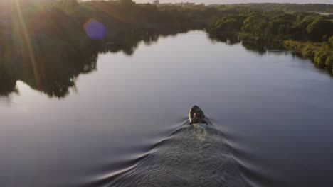 Inflatable-pontoon-boat-drives-up-river-as-sun-flare-shines-down-and-wake-breaks-calm-water-surface