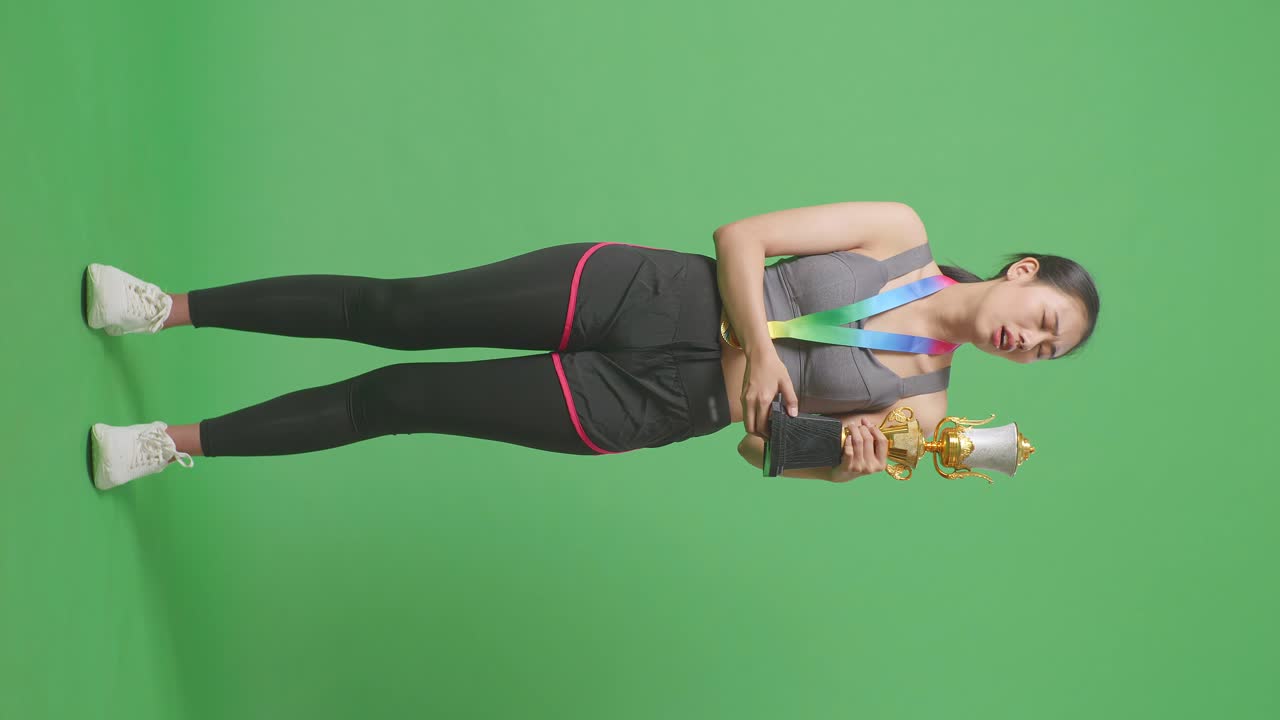Donna asiatica a figura intera, contrariata, con una medaglia d'oro, che guarda il trofeo e poi scuote la testa, insoddisfatta della vittoria come prima classificata su sfondo green screen in studio.