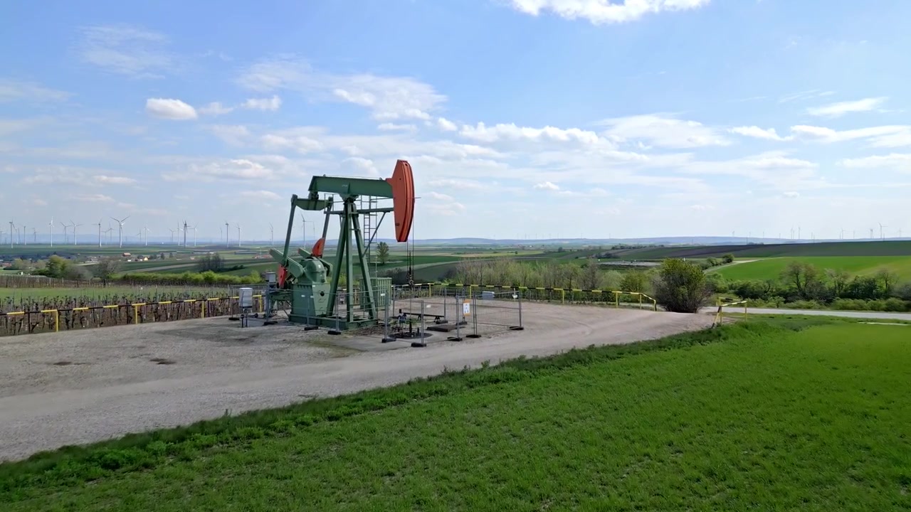 Drone Approach Towards Pump Jack Extracting Gas From Oil Field. Wind Farm In Distance. low aerial