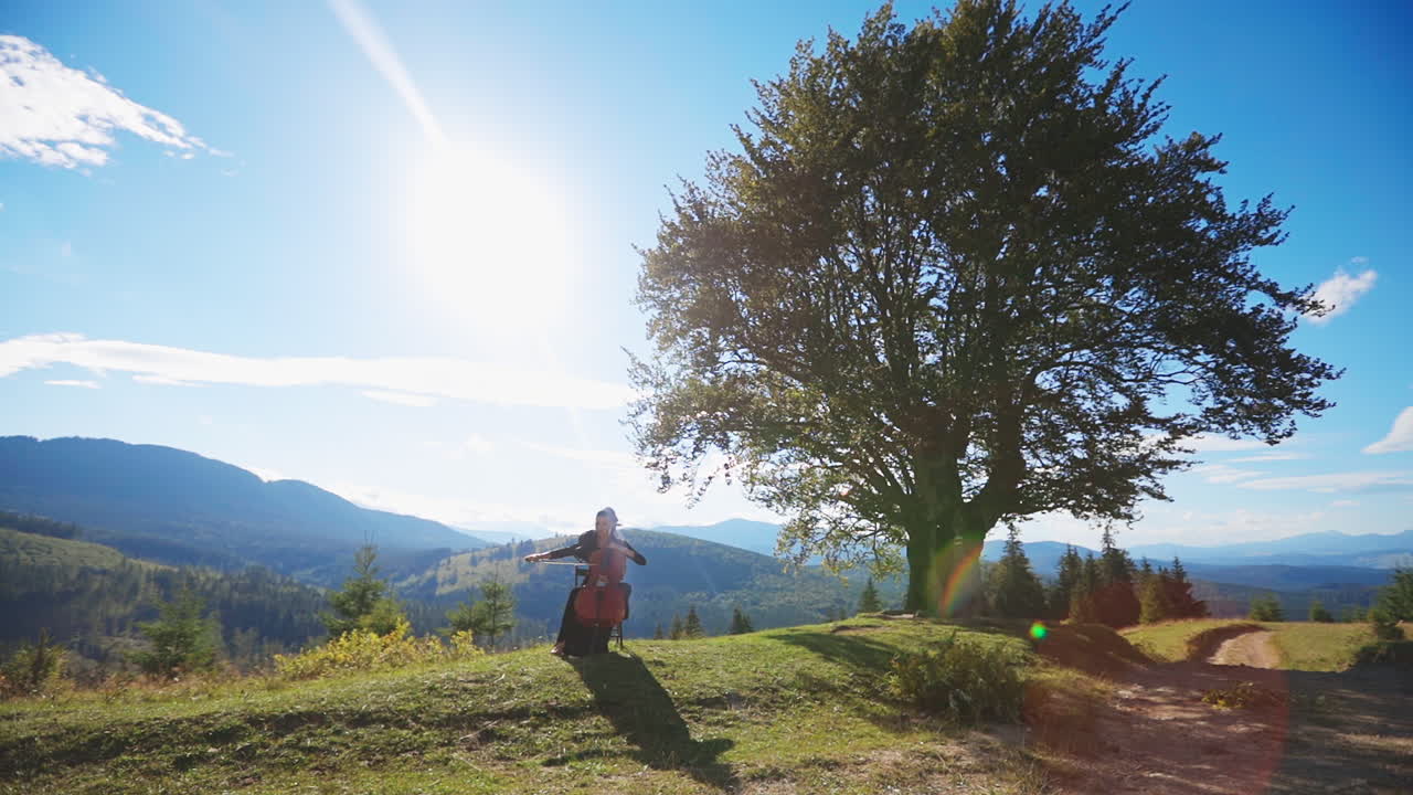 Woman playing cello on the hill on wonderful sunny day. Performing classical music at the backdrop of mountains on sunny day.