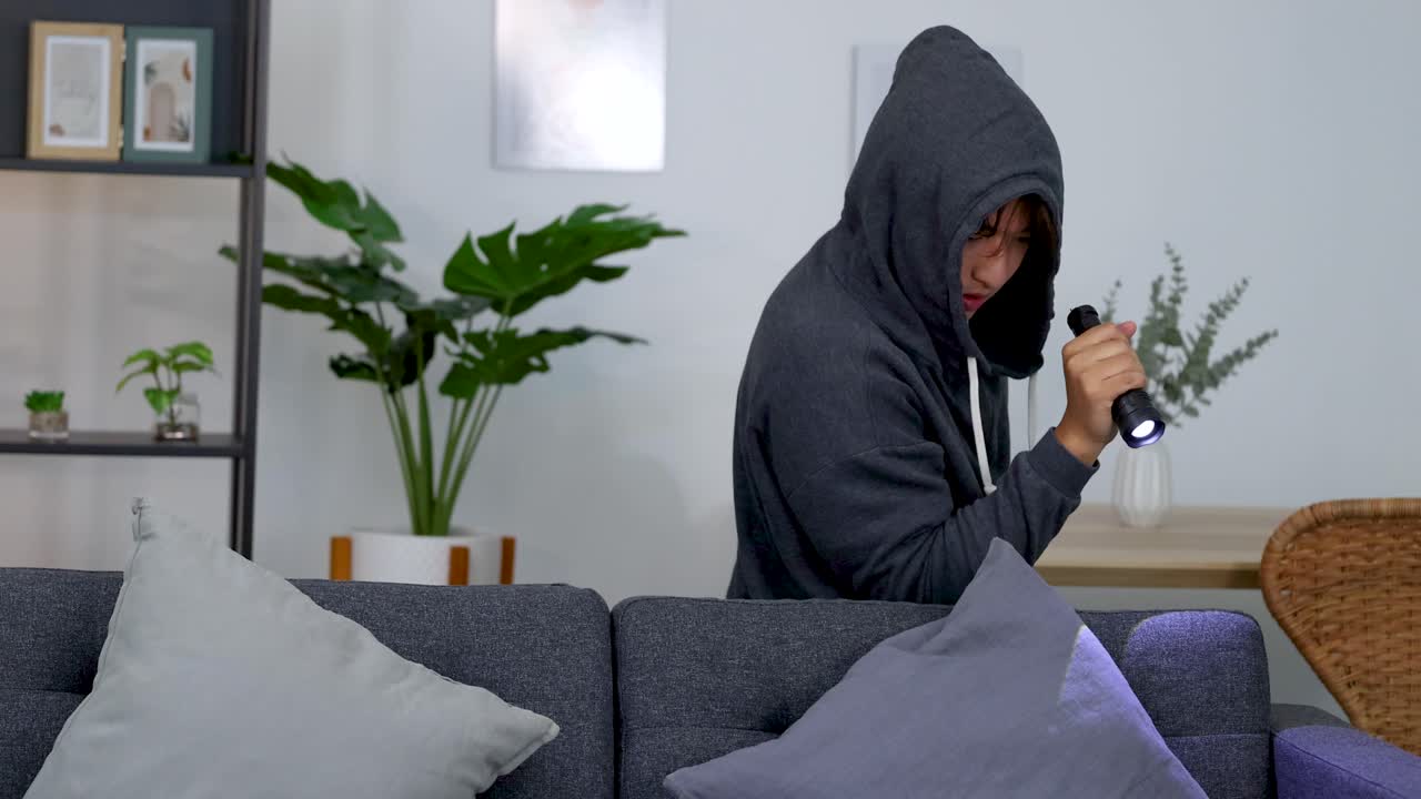 A hooded burglar cautiously searches a modern living room using a flashlight during a robbery
