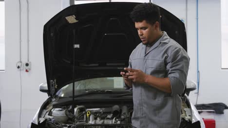 african american male car mechanic looking at an open car engine and talking on a smartphone