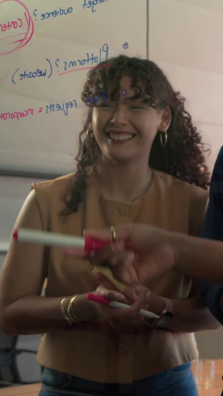 Video verticale: Donna con top color cuoio che prende un pennarello da destra, sistema post-it su una lavagna bianca, pianificando.