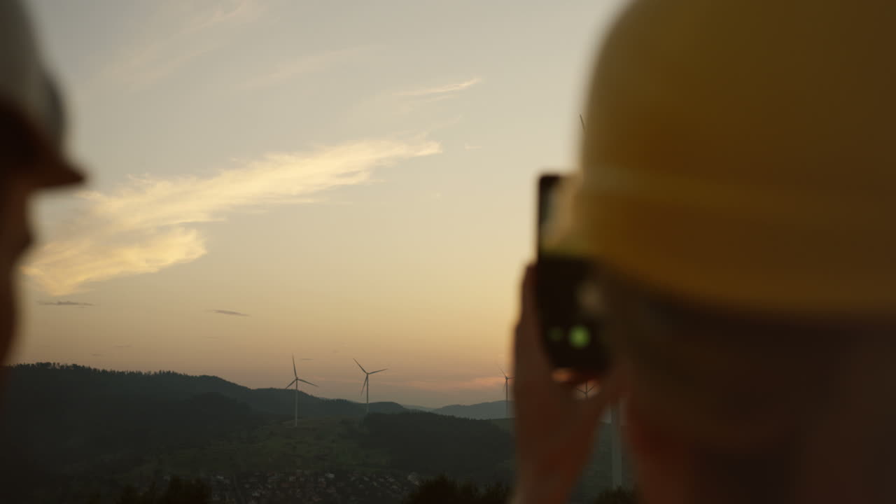 vista trasera de una trabajadora caucásica usando un casco tomando una foto con su teléfono inteligente de las turbinas de los molinos de viento girando al atardecer