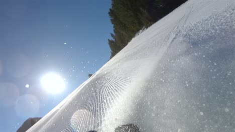 a skier going down a steep slope, in the background a blue sky with bright sun and snow rising from the skis view from the skier’s back