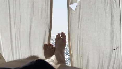 person rests with feet up, framed by light curtains and ocean in background. serene seaside moment, sunlit waves and gentle breeze create a tranquil vacation mood