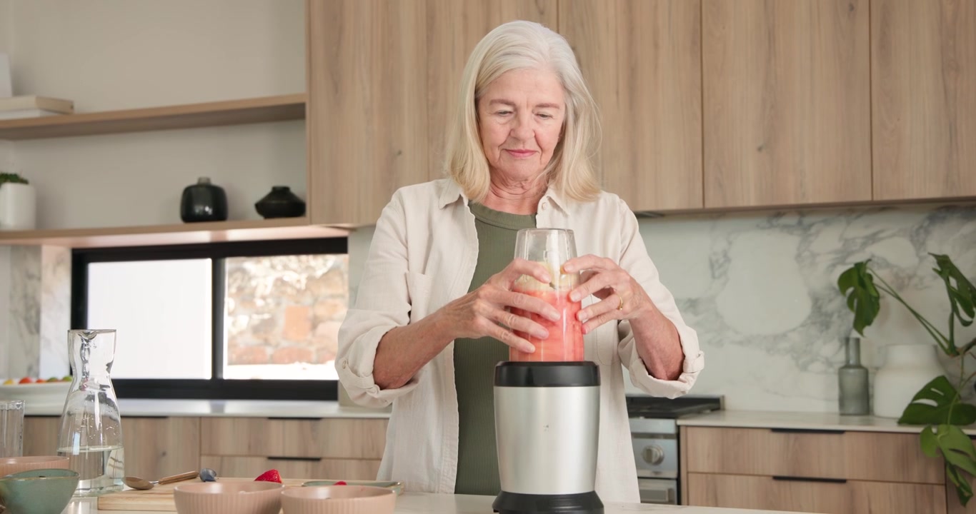 Una donna anziana versa acqua in una tazza, poi sigilla e blocca il coperchio sulla base del frullatore per preparare un frullato per il pasto.