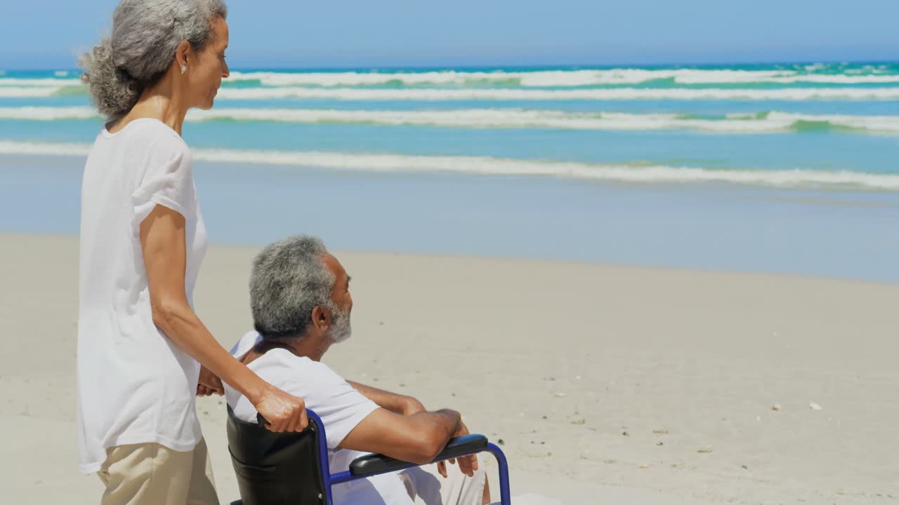Side view of active senior African American woman with disabled senior man on beach in sunshine 4k
