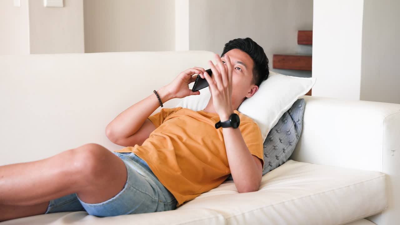 Relaxing at home, young man talking on smartphone while lying on couch
