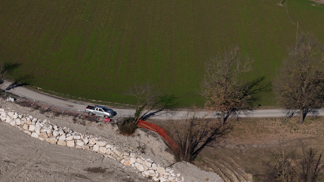 Drone footage captures a silver pickup truck navigating a winding road through a scenic rural landscape with green fields and bare trees