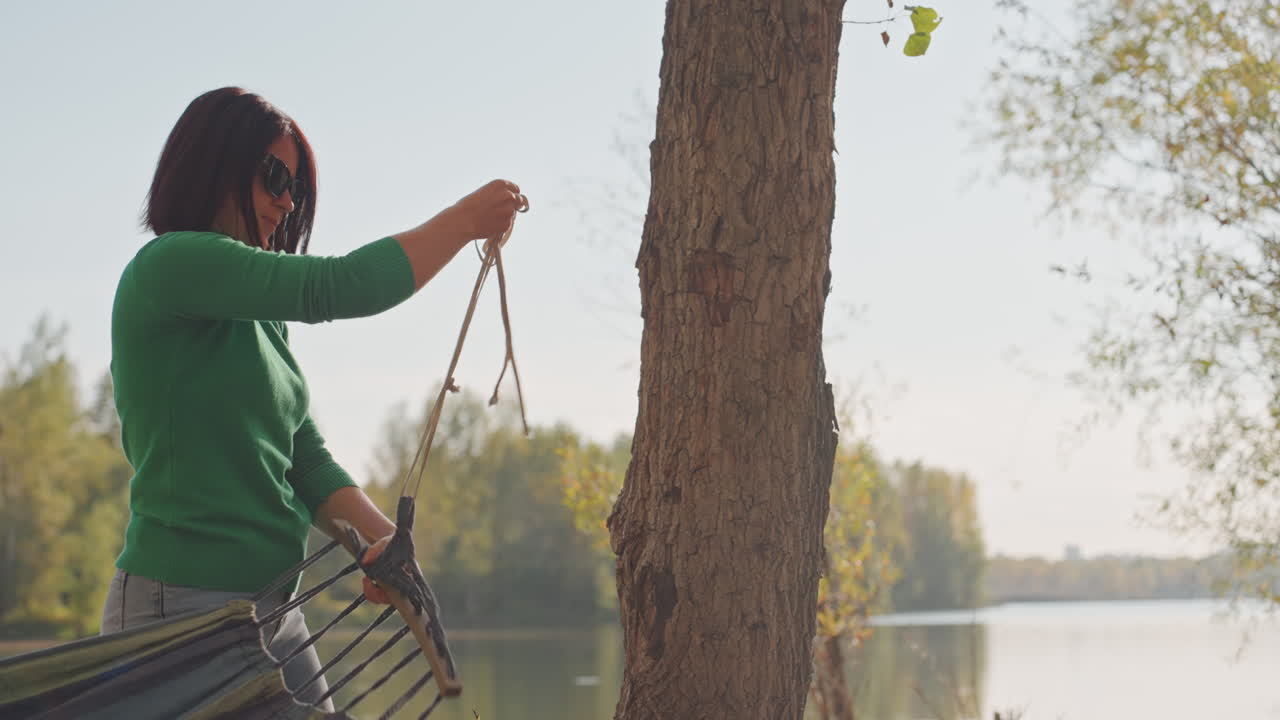 Caucasian woman attaching hammock between trees, green sweater and sunglasses, securing ropes to trunk, calm lake and treeline backdrop, hands tying knot, gentle sunlight, relaxed lakeside camping