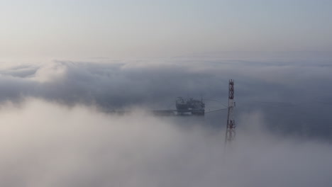 fly over a dense cloud of fog, covering a telecommunication tower,standing on the hill, with oil terminal and oil tanker moored far in the distance