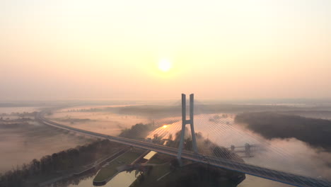 Toma-Aérea-De-Un-Moderno-Puente-De-Carretera-Por-Cable,-Sobre-Un-Amplio-Río-Tranquilo,-Durante-Un-Amanecer-Brumoso