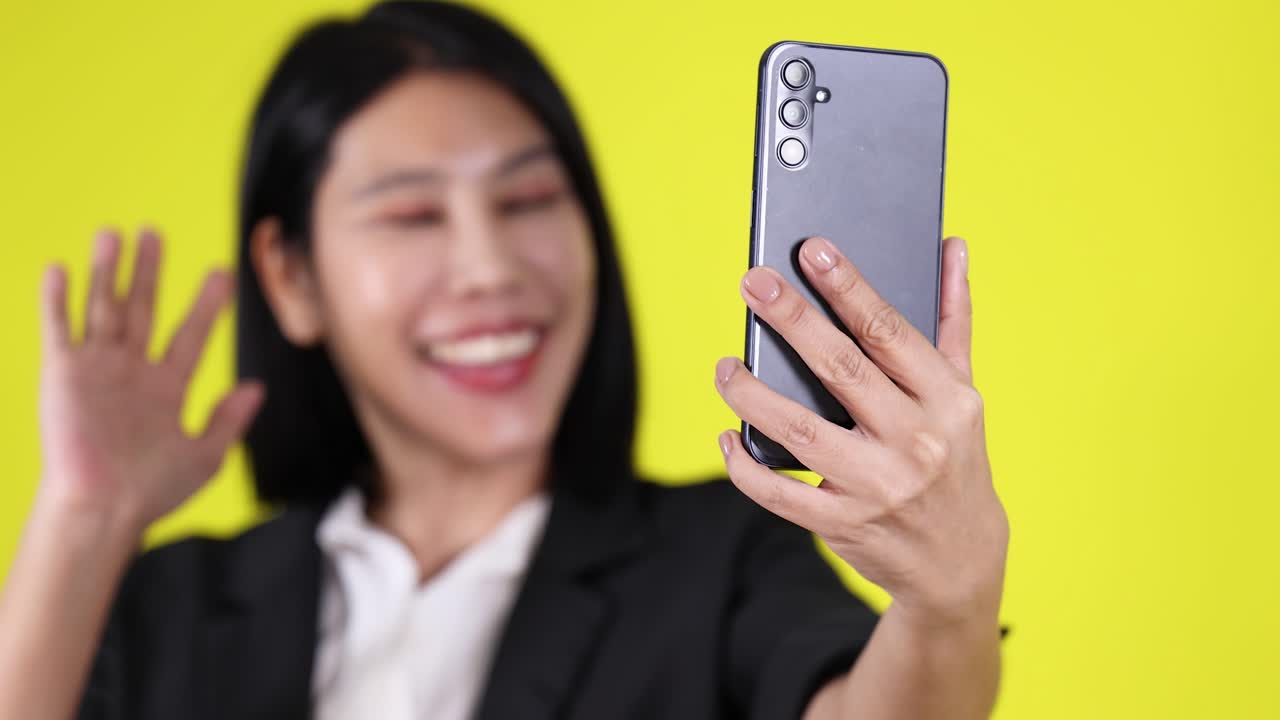 Confident woman in black blazer waves and smiles on video call, yellow background, soft lighting