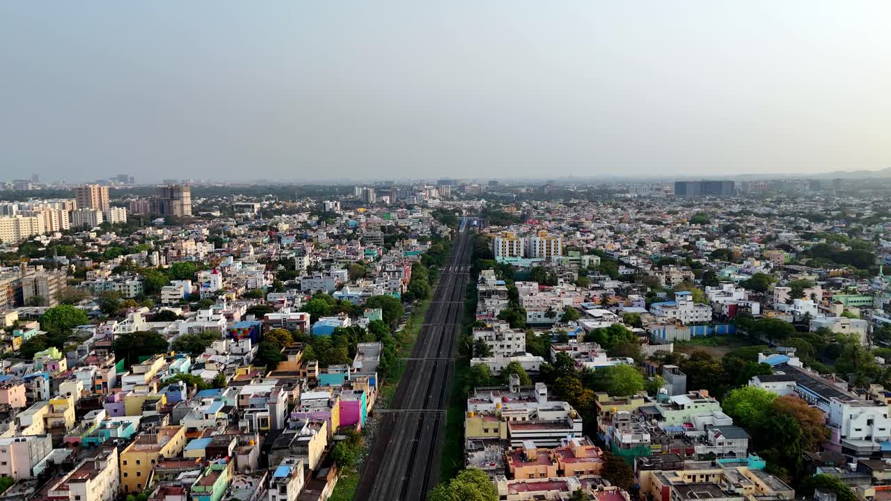 This aerial footage of Chennai's Saidapet depicts a vibrant urban tapestry. Densely packed homes fill the foreground, with a prominent railway line to the right. Modern high-rise buildings,
