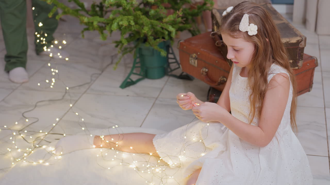 Young girl in white dress sitting on floor and arranging Christmas lights, a festive atmosphere with a Christmas tree and cozy home setting, showcasing holiday preparations