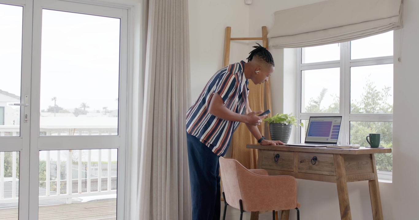 Homem afro-americano verificando smartphone, caminhando até a secretária em casa, comparando telefone com laptop.