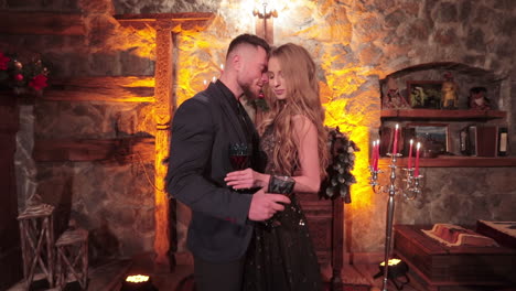 beautifully dressed young people standing in the nice room with stone walls. couple is holding filled glasses and hugging sweetly.