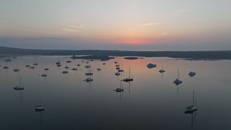 Pink-blue-sky-reflecting-in-calm-waters-with-sailboats,-Fornells-Bay