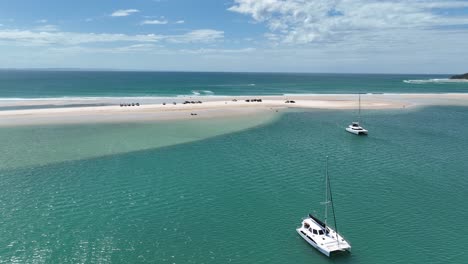 flying over catamaran in bay towards 4x4 vehicles parked on white sandy beach