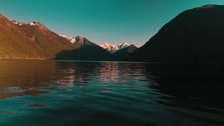 Acque profonde, scure e incontaminate del lago che increspano dolcemente, estendendosi verso spettacolari e aspre montagne alpine con chiazze di neve bianca brillante che risplendono alla luce dorata del sole serale, riprese da un volo aereo a bassa quota.