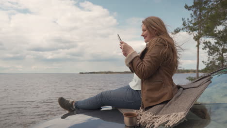 Red-Haired-Woman-Sitting-On-Her-Car,-She-Takes-A-Picture-Of-The-Beautiful-Landscape-Of-The-Lake-In-Which-She-Is