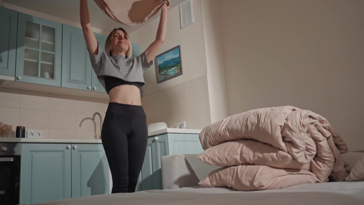 Donna bianca che balla mentre rifà il letto, amante della musica che si muove al ritmo di una playlist vivace nella cucina di un monolocale luminoso, lanciando un piumino beige con movimento energico, sorridente in crop top e leggings, piegato.