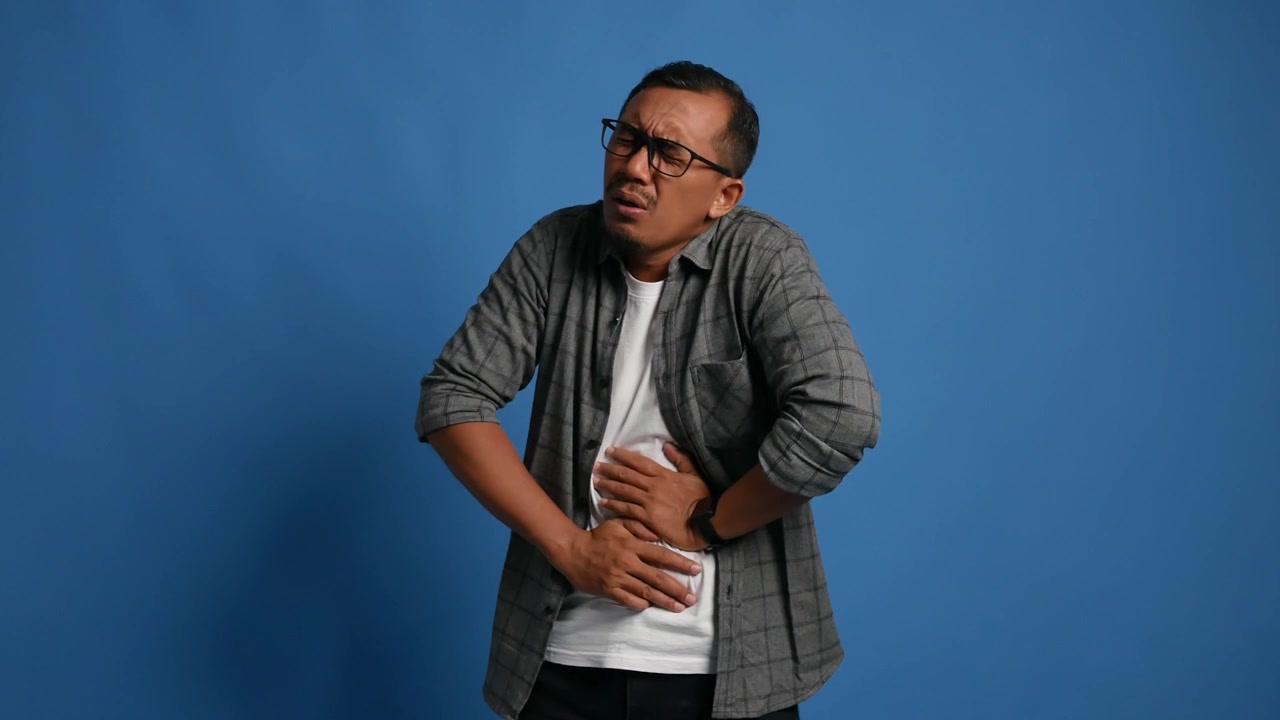 Adult man standing isolated on blue background, clutching stomach due to pain.