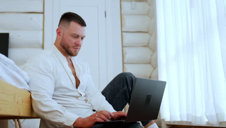 man in white shirt sits near the bed working on laptop. low angle view at the freelancer using computer for work remote.