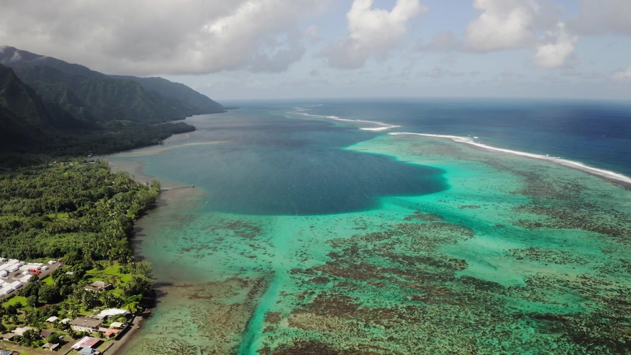 Un drone aérien survole un trou bleu turquoise dans le récif corallien de Teahupoo, Tahiti, avec des sommets de vallée brumeux et un village côtier encadrant la scène au loin.