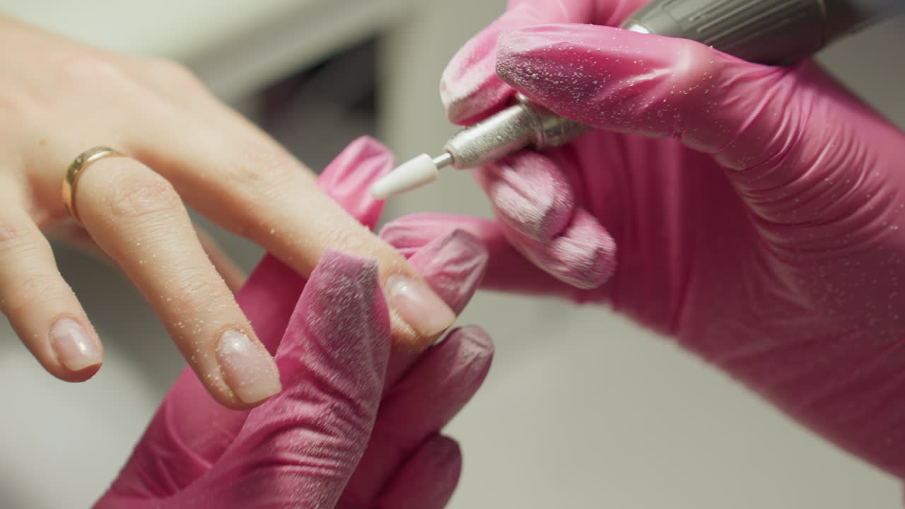 Close-up of nail technician in pink gloves carefully filing client nail using electric tool, with dust particles covering gloves and technician moves to next finger during manicure process