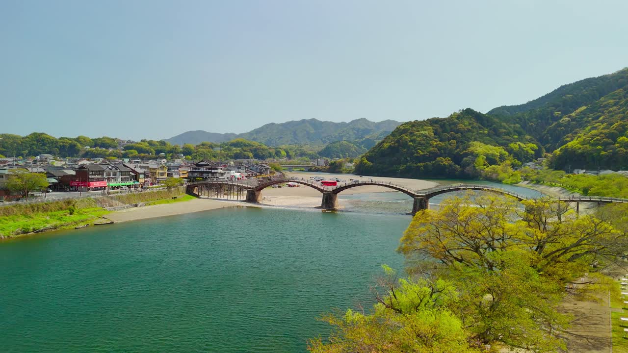 A beautiful aerial shot pans across the historic Kintai Bridge, a wooden arch bridge spanning the Nishiki River in Iwakuni, Japan. The scene captures the picturesque town, lush green mountains